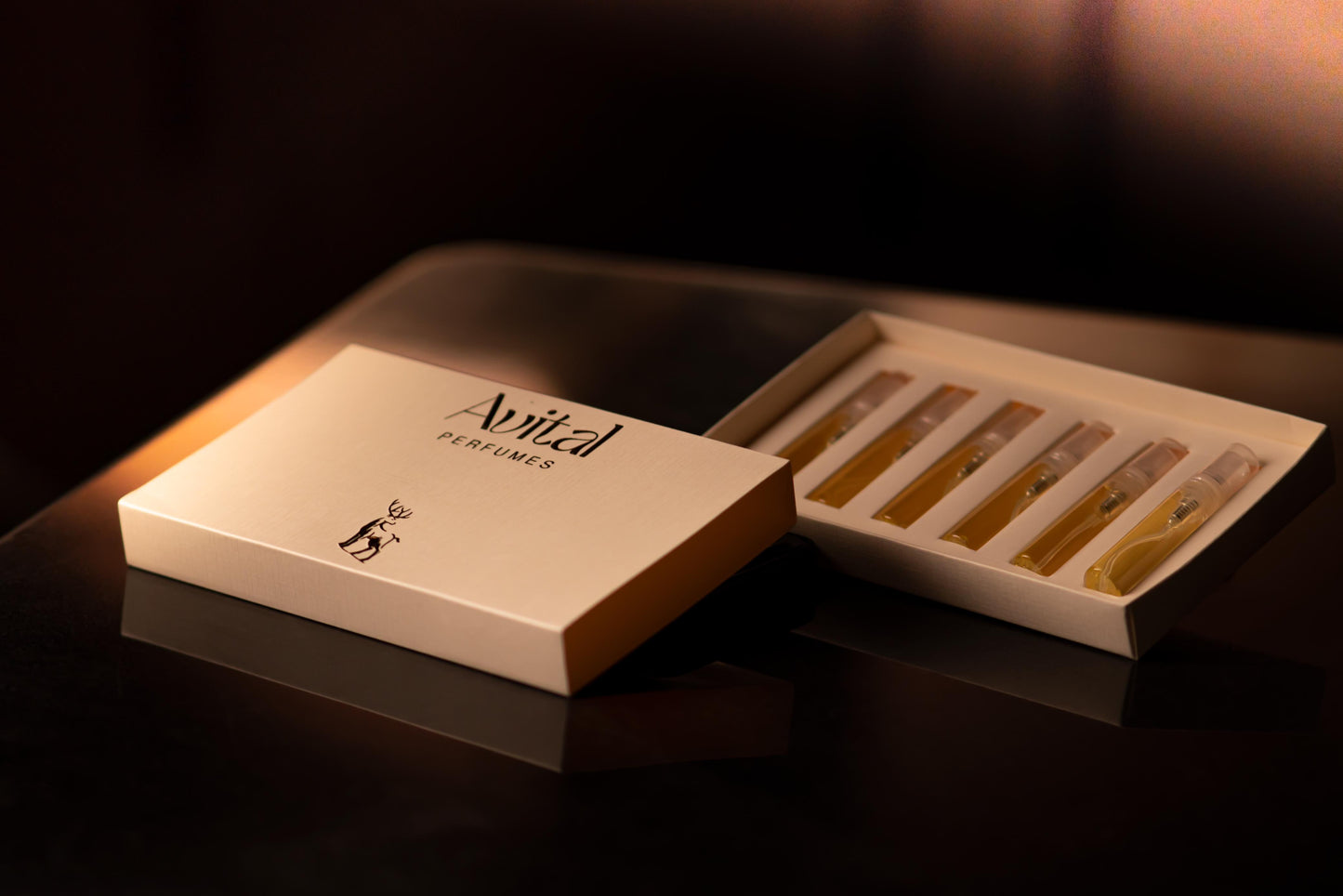 An elegant, cream-colored box labeled 'Avital Perfumes' is partially open to display six 5ml testers of perfume. The testers are arranged in a single row and filled with perfumes. The box features black text and a small deer logo, all highlighted by warm, ambient lighting that enhances its luxurious and refined presentation